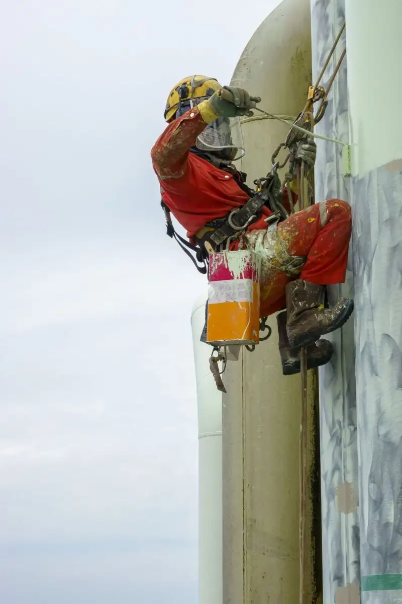 Alpinist utilitar care vopsește un coș industrial, cu găleata prinsă de ham
