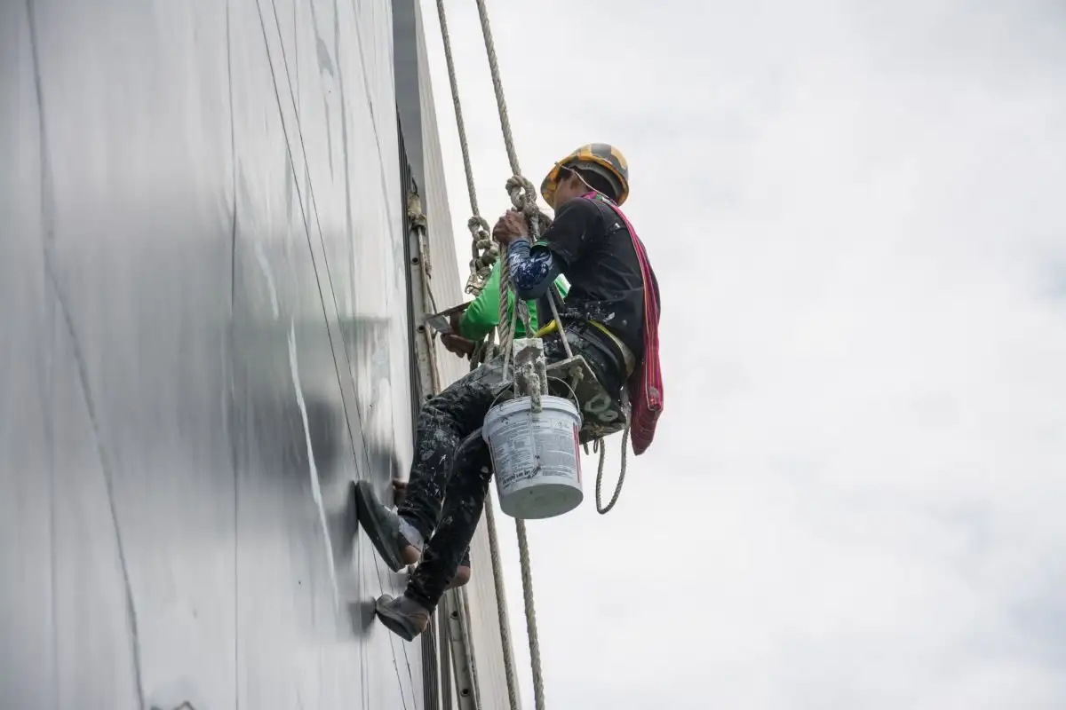 Alpinist utilitar care vopsește o fațadă, cu găleata prinsă de ham