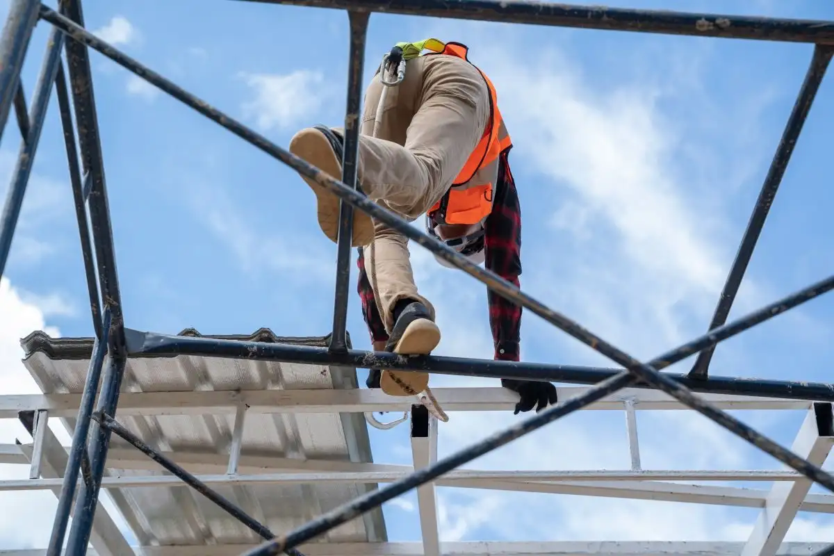 Lucrător care traversează o structură din grinzi metalice, văzut de jos