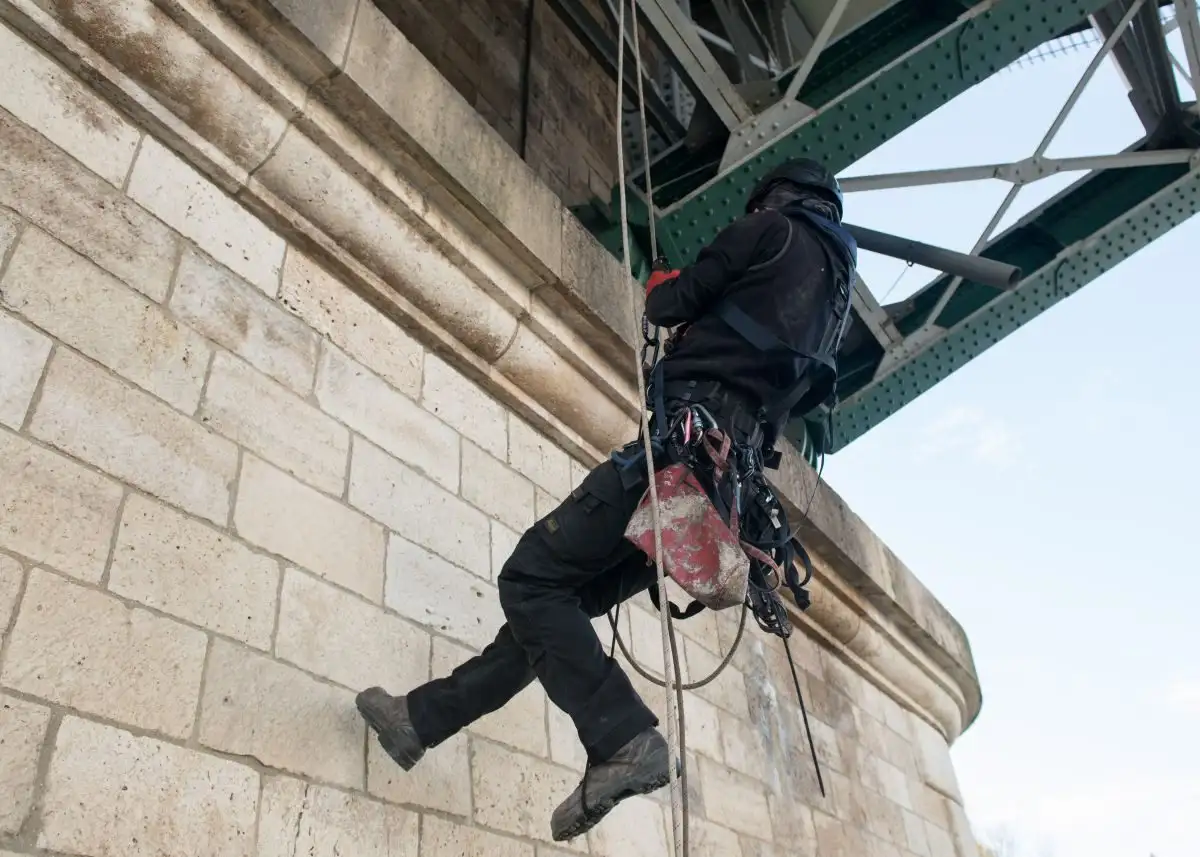 Alpinist utilitar pe corzi la un pilon de piatră, sub un pod metalic