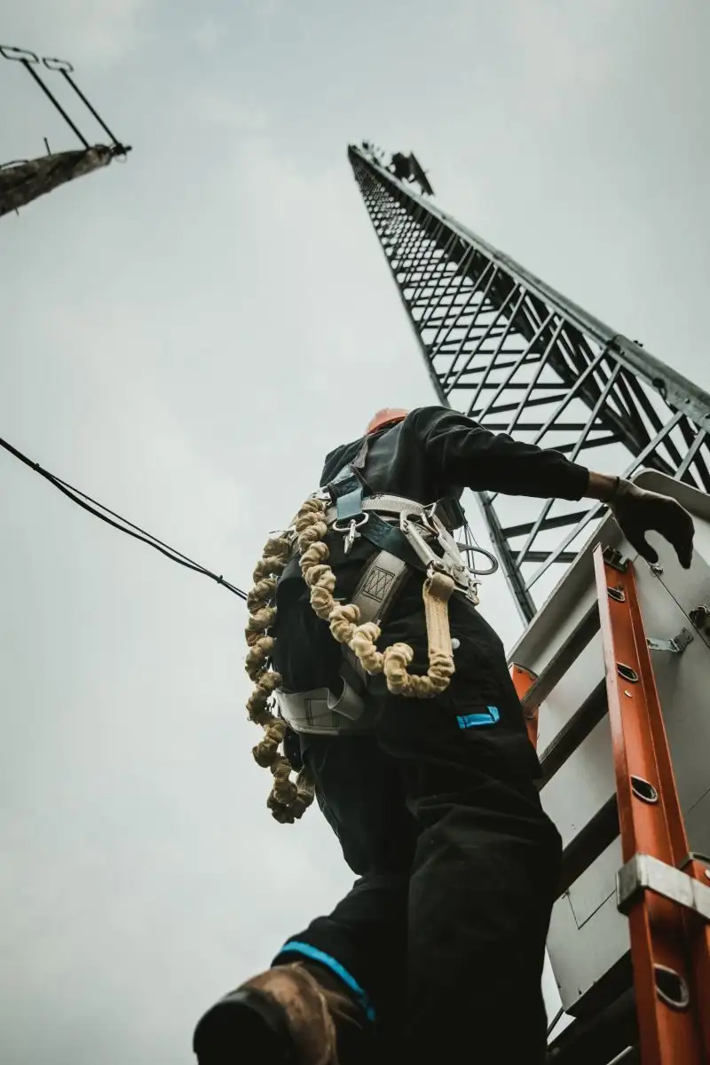Lucrător care urcă pe scara unui turn metalic, asigurat cu coardă